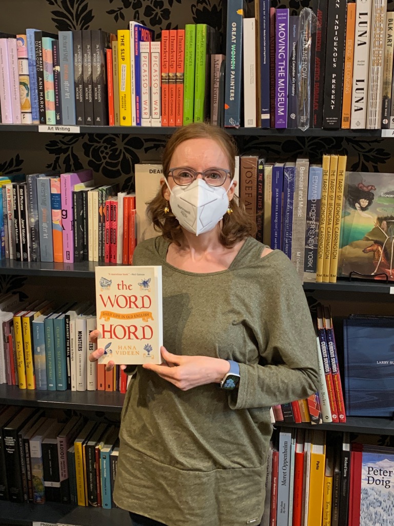 In a bookstore the author, a white woman with red hair, glasses and face mask, holds a paperback copy of “The Wordhord: Daily Life in Old English” by Hana Videen.