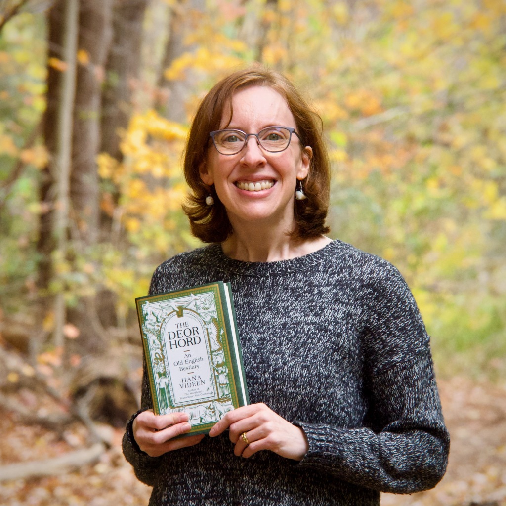A red-haired, glasses-wearing, thirty-something woman with bird earrings and a big smile holds a green and gold book, "The Deorhord: An Old English Bestiary" by Hana Videen; the background is a forest with autumn colours.